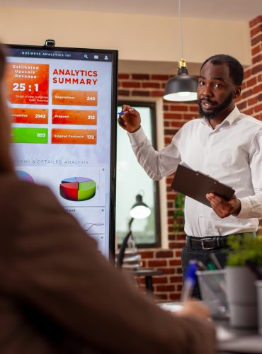 Businessman with clipboard in hand, using digital monitor to explain analytics summary in meeting. African american entrepreneur doing project presentation, planning marketing strategy with his team.